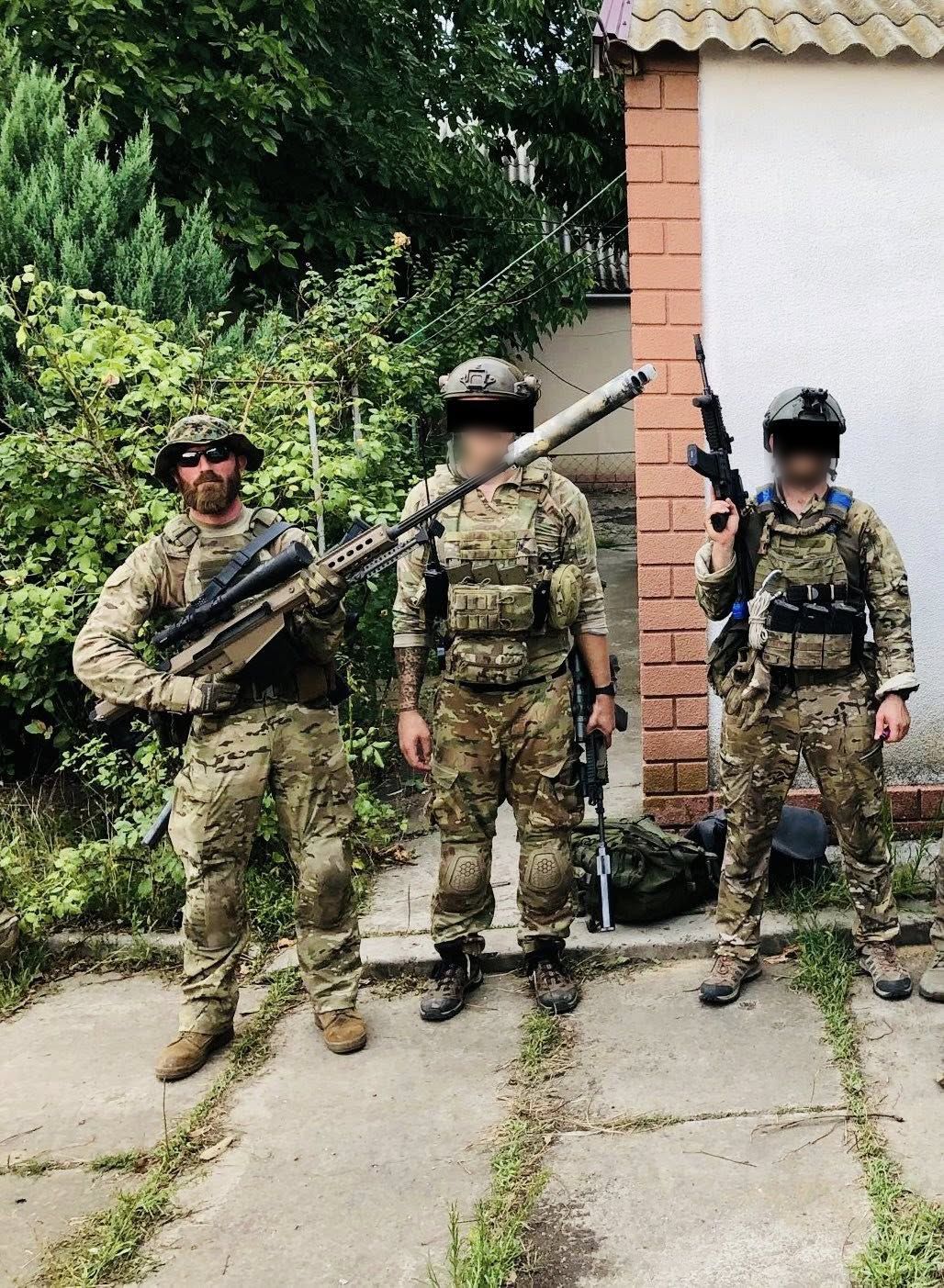 Three operators in multicam combat fatigues with rifles standing in a yard; two of the three faces are redacted.
